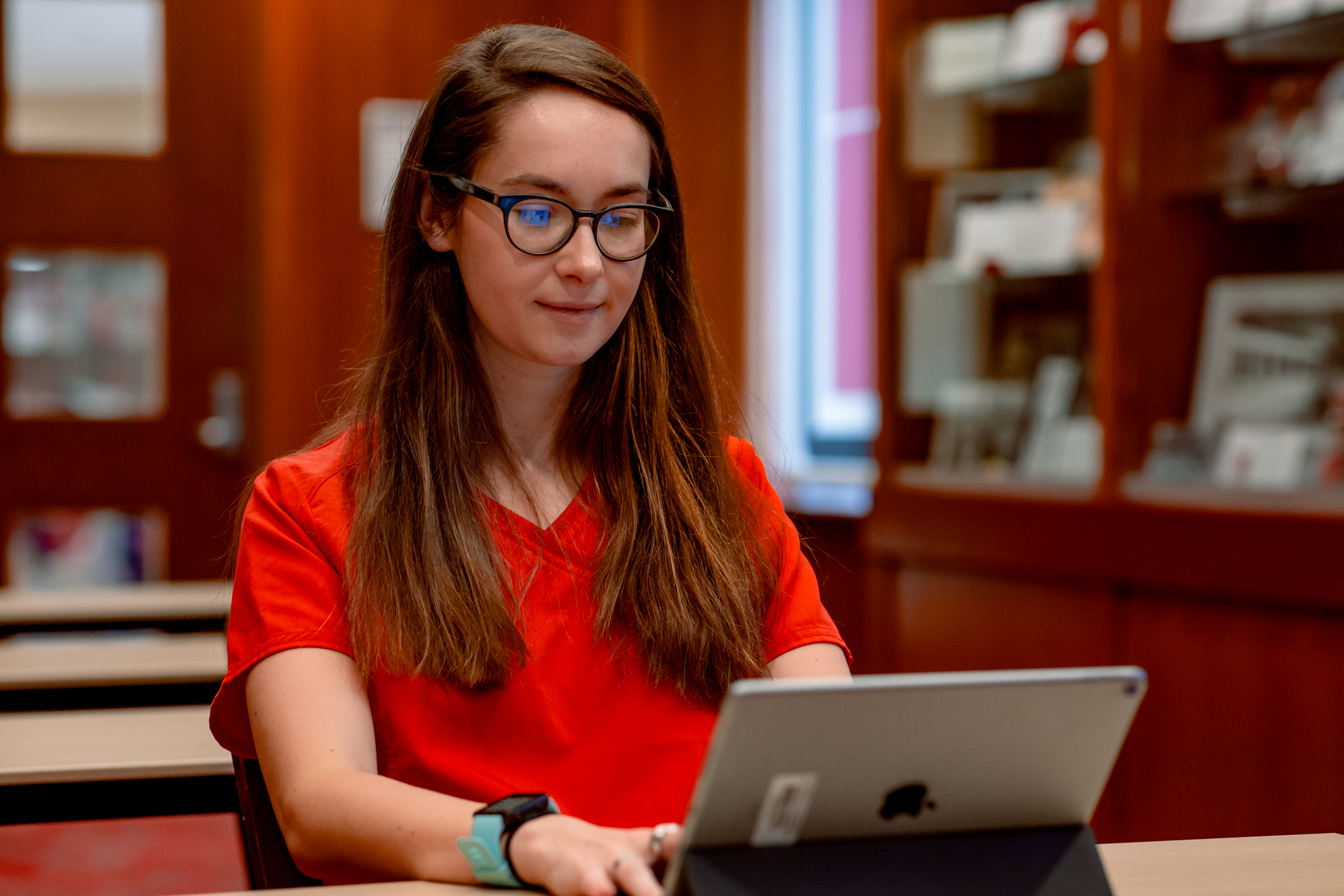 A nursing student in red scrubs completes work for her online nursing program on a tablet.