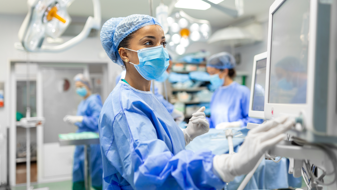 A black female nurse anesthetist working in a surgical room wearing protective gear checking monitors while sedating patient before surgical procedure.