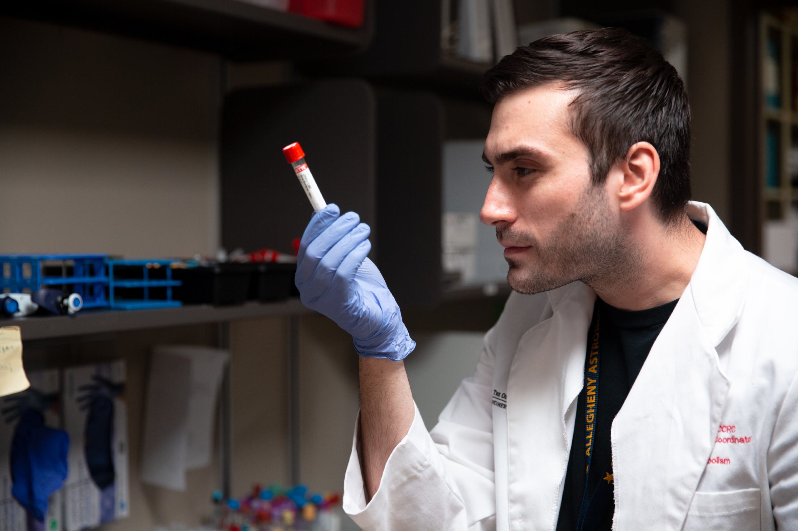 A young man in a lab examines a vial closely while wearing a glove. 