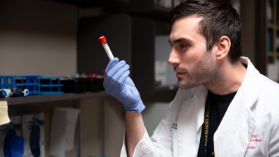 A young man in a lab examines a vial closely while wearing a glove. 