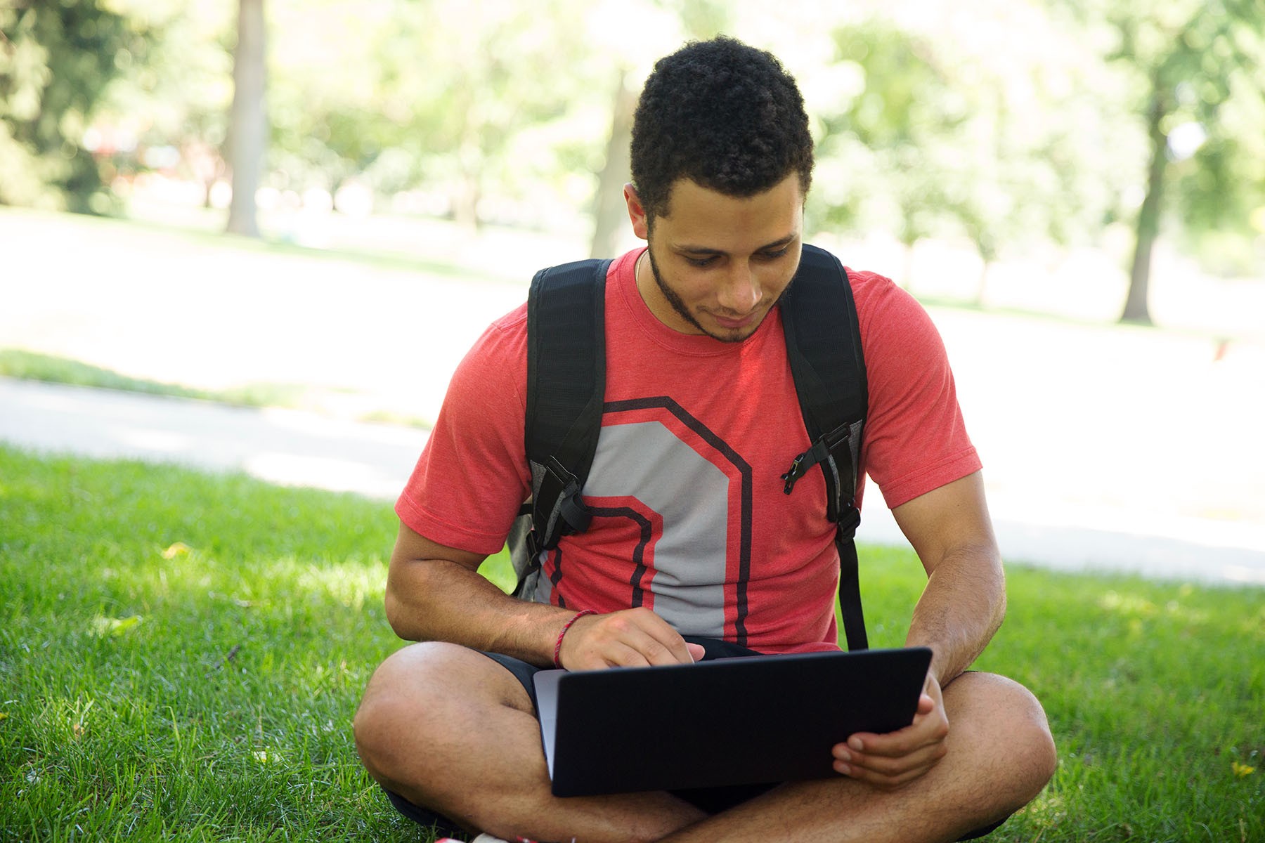 Young man wearing a red Ohio State T-shirt sits on the grass outside while looking at a laptop