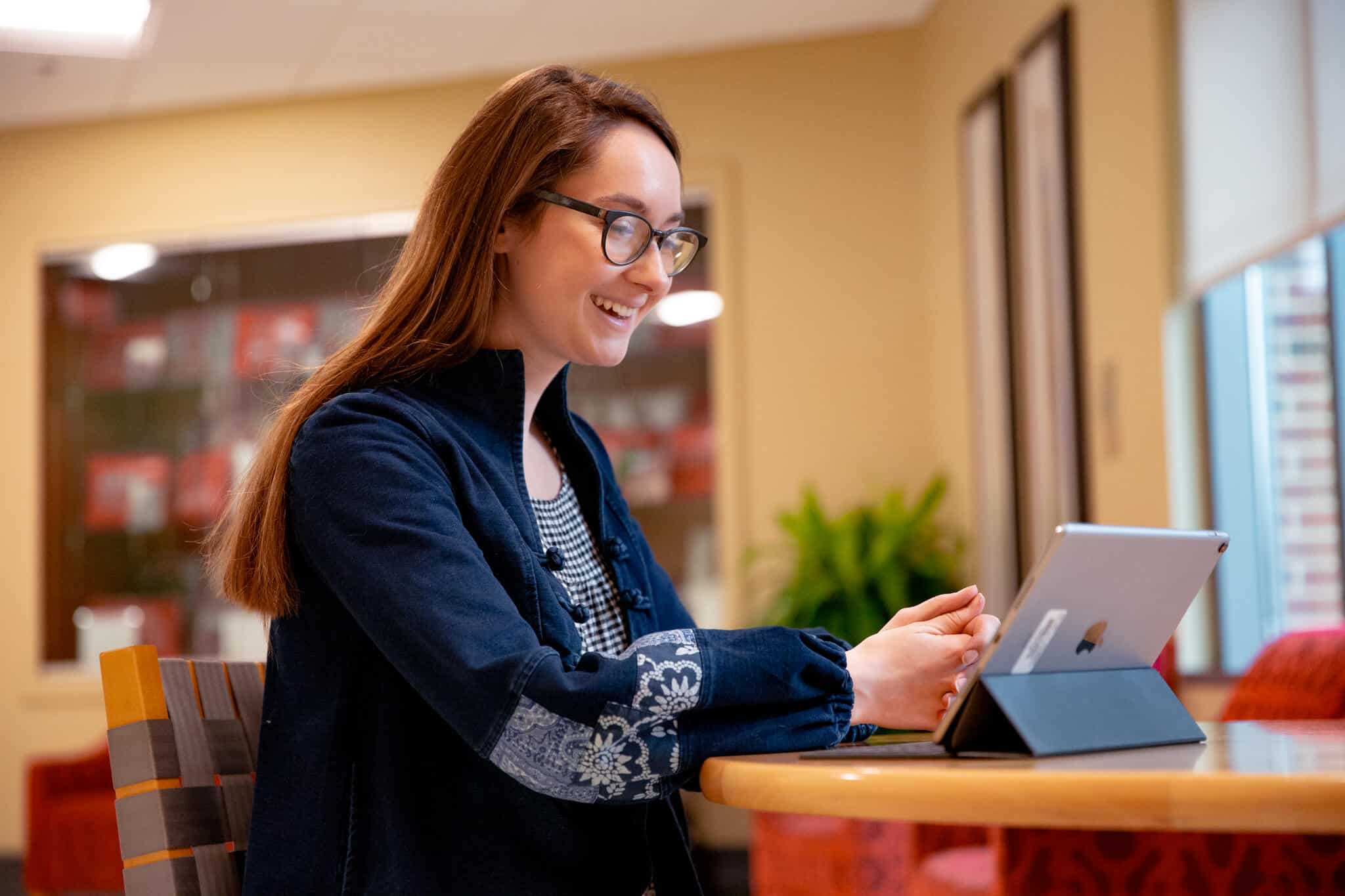 A student sits in a library smiling while working on her studies on an iPad.