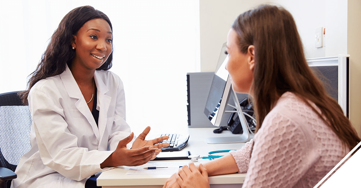 A doctor wearing a white coat sits at a desk talking with a patient.