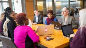 A group sits with laptops around a table in a workspace