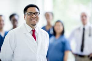 Man with DNP degree in a white coat and red tie stands proudly amongst peers.