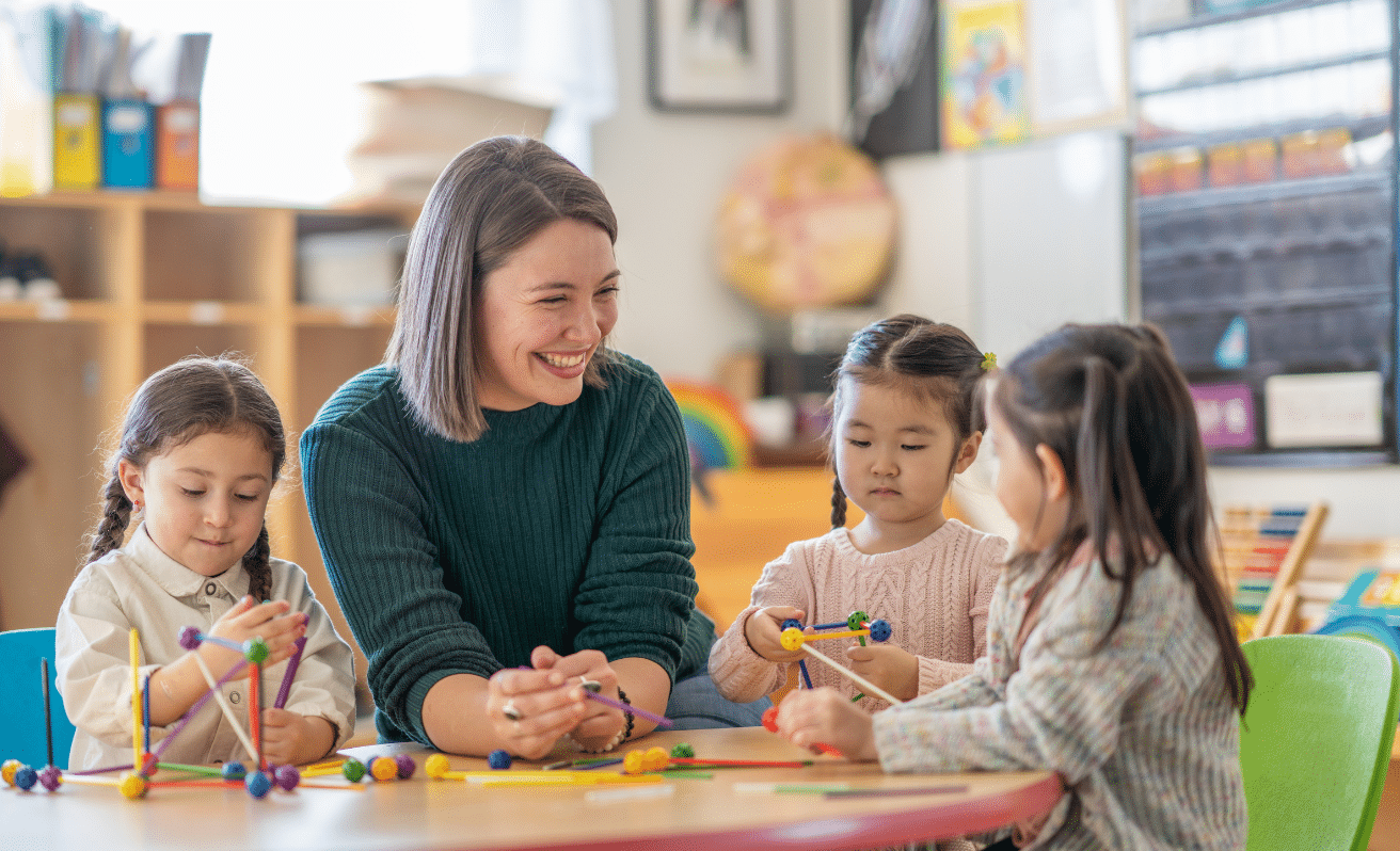 A pre-school teacher smiles and sits with 3 students in a classroom.