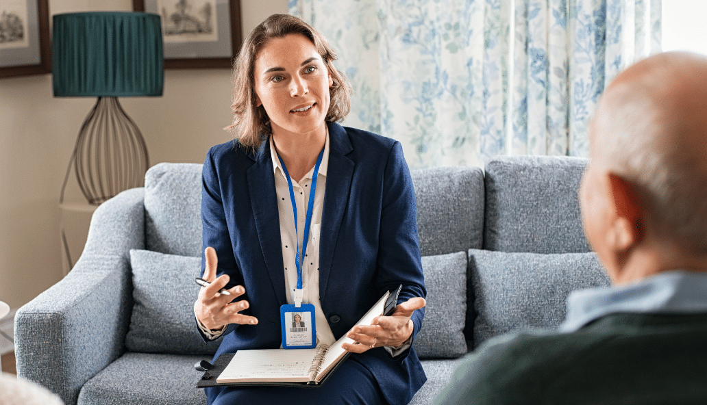 A professional woman with an open notebook sits on a couch consulting with an elderly couple.