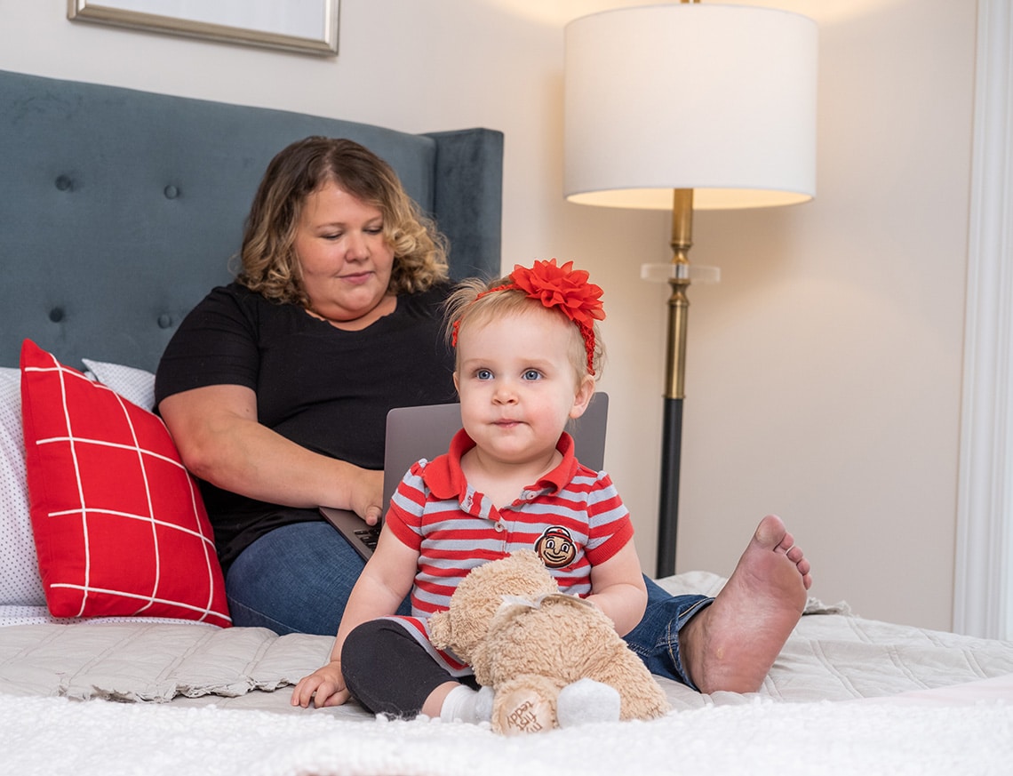 A mother working on her laptop while sitting on her bed with her infant daughter.