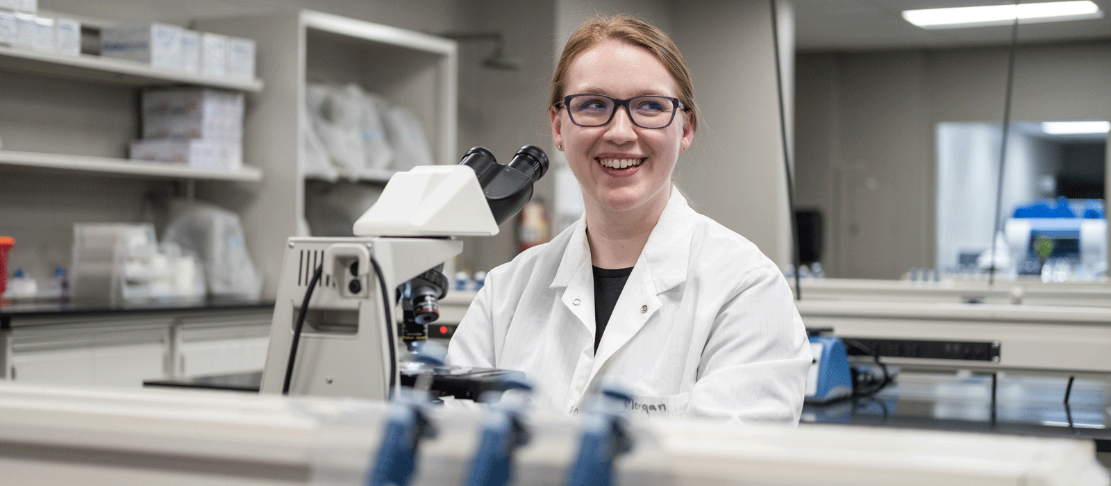 Morgan Cummings in the lab standing at a microscope smiling.