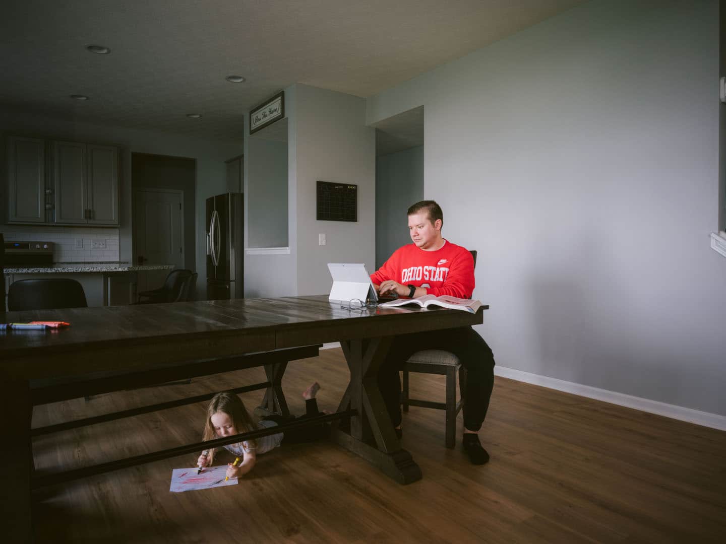 Man sitting down at table looking at laptop while kid plays underneath table.