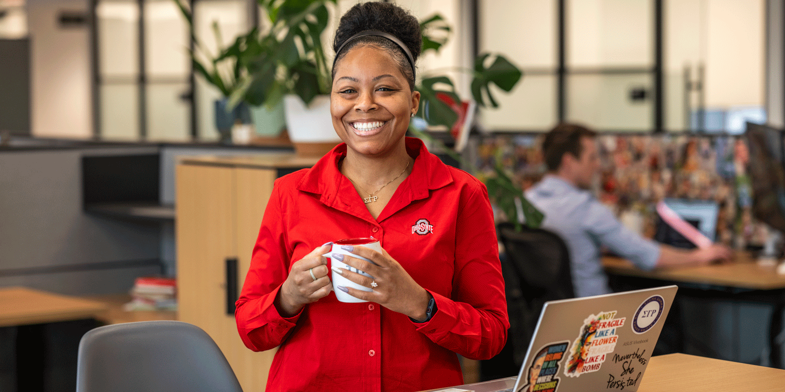 Photo of Ari holding a coffee cup in an office next to her laptop.