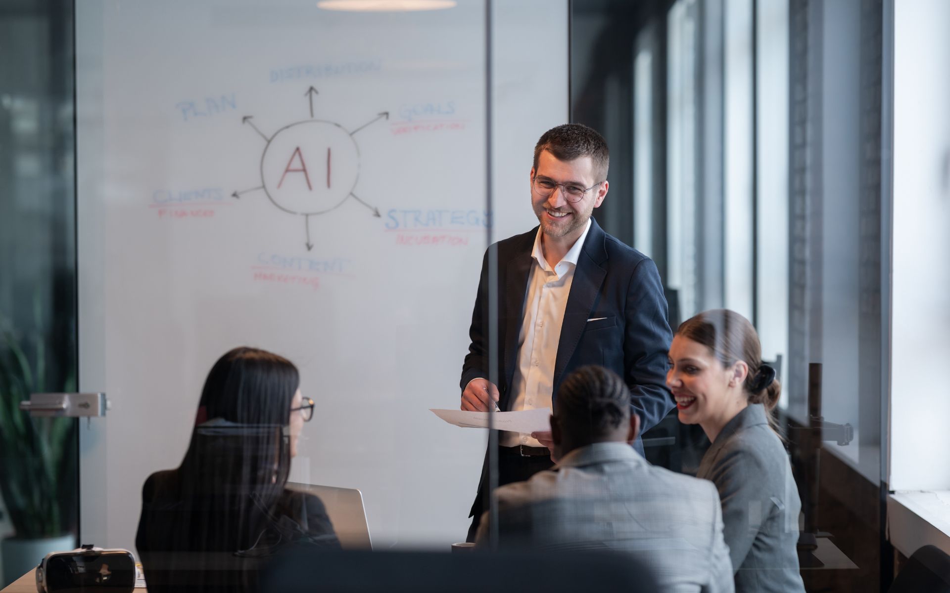 A man in a suit smiling while presenting to a group of people in a modern office with an AI diagram on a glass whiteboard.